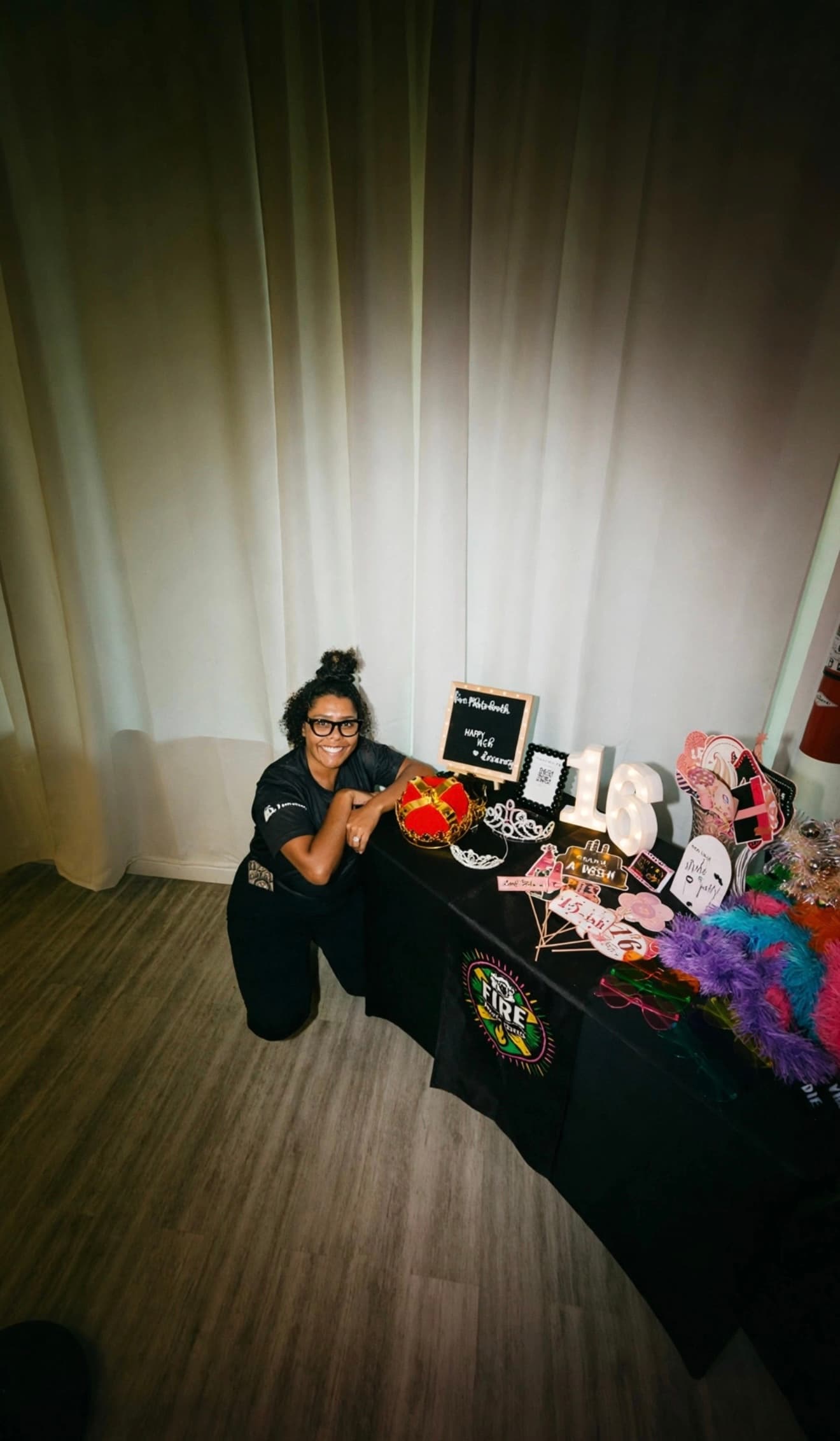 Smiling woman kneeling beside a table of 16th birthday party props and colorful feather boas.