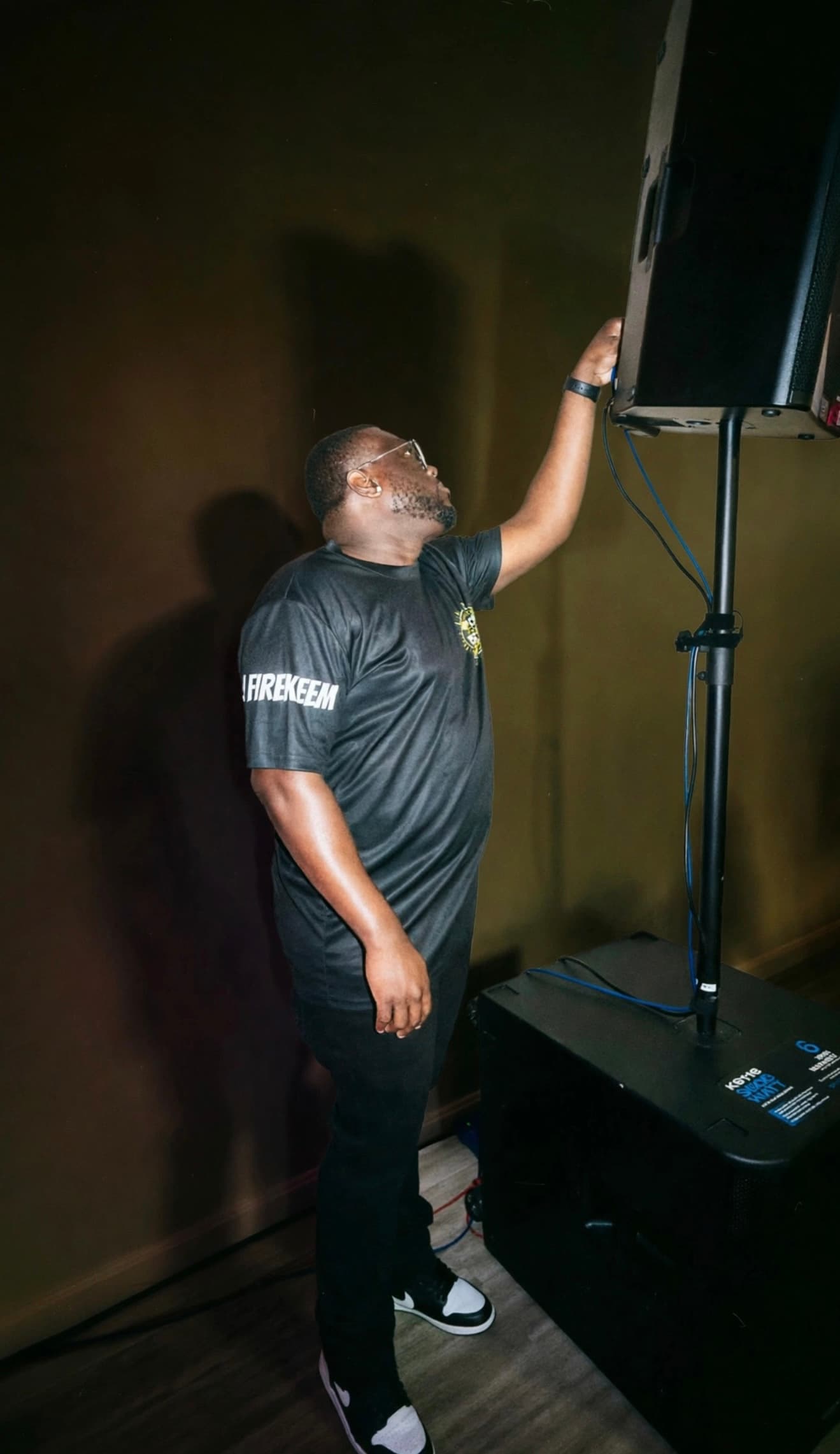 Man in black t-shirt adjusts a speaker mounted on a stand above a large subwoofer.