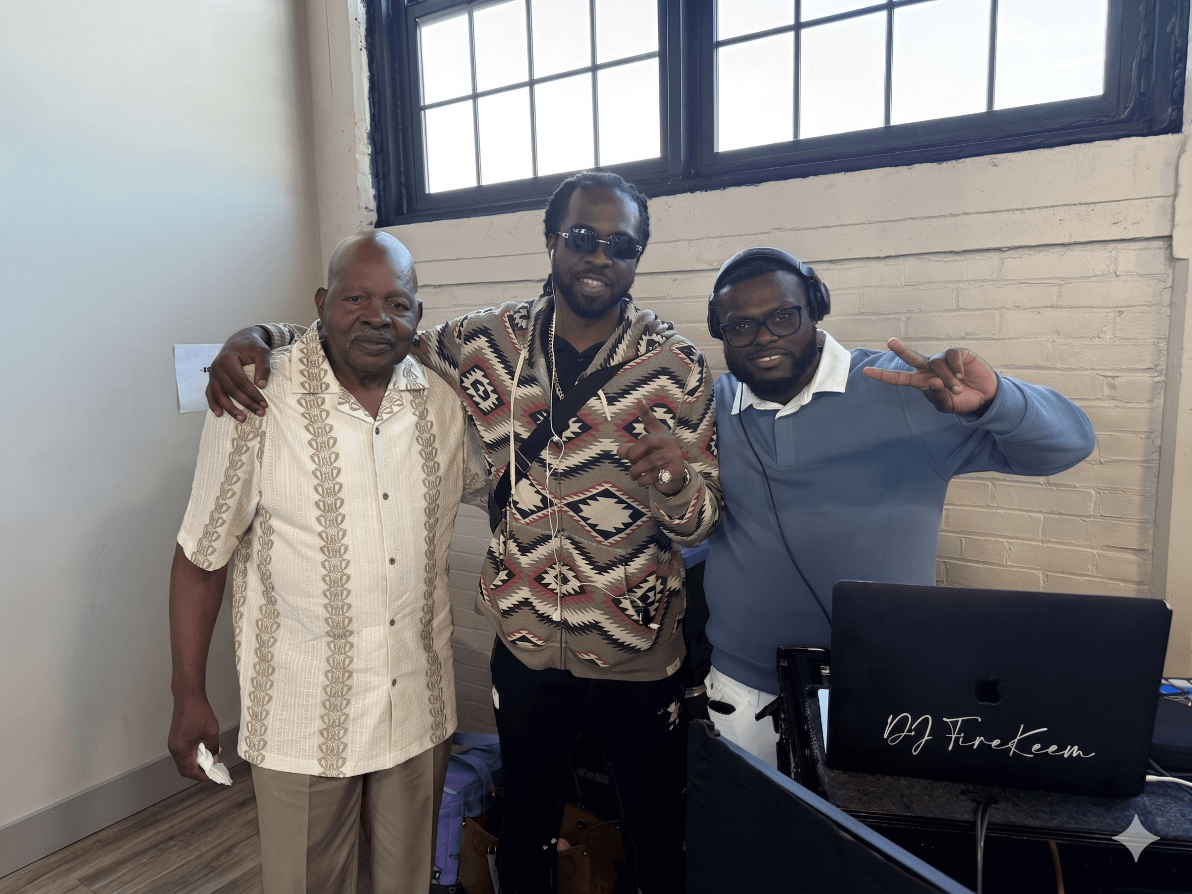 Three Black men pose together indoors, one wearing headphones beside a DJ FireKeem laptop.