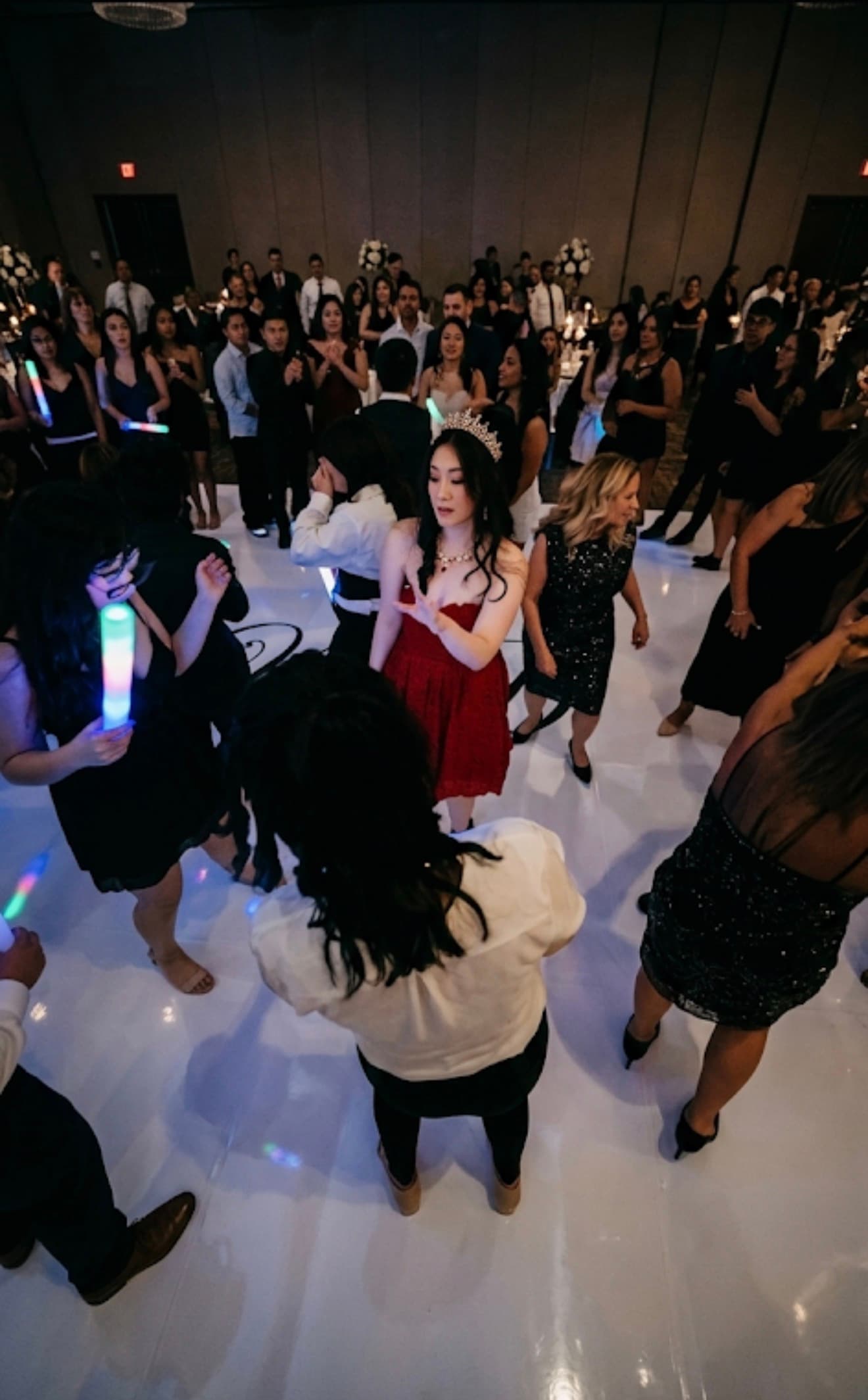 Woman in red dress and tiara dances on white floor among guests holding glowing light sticks.
