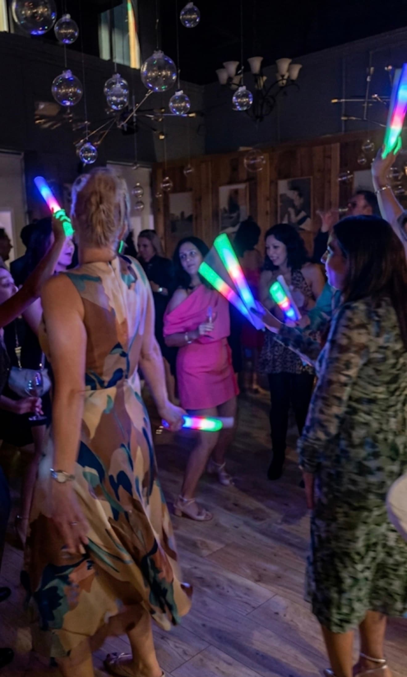 People dancing at a party waving colorful LED foam sticks under hanging glass globes.