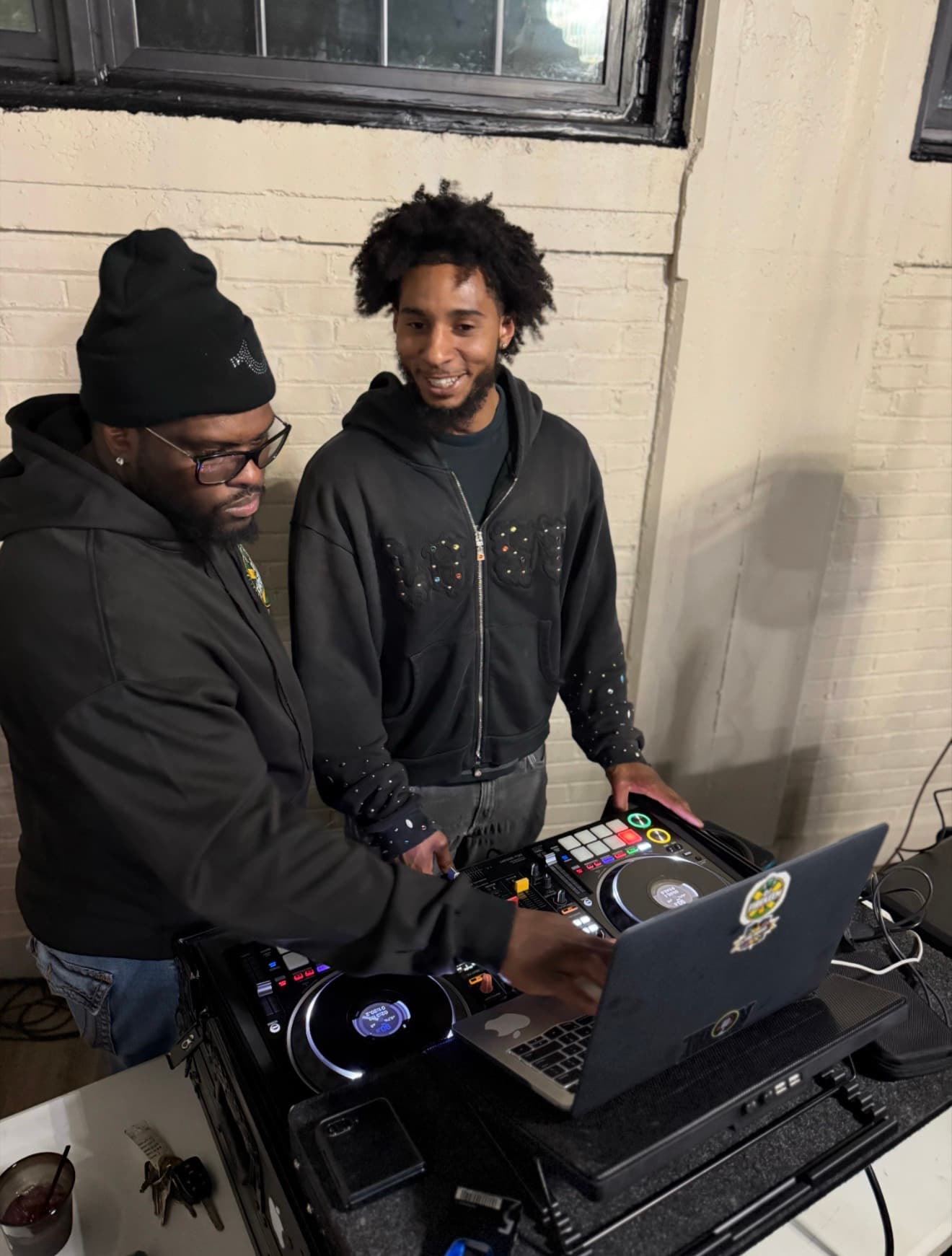 Two men in black hoodies operate a DJ controller and laptop against a brick wall.