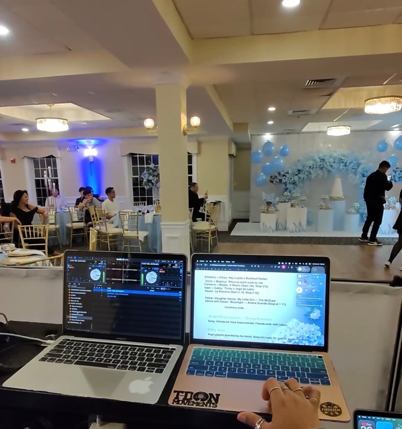 DJ booth with two laptops overlooking a banquet hall decorated with blue balloons and flowers.