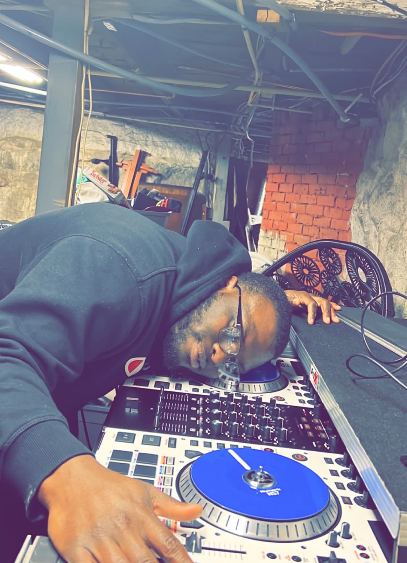 Man in a black hoodie and glasses resting his head on a DJ controller.