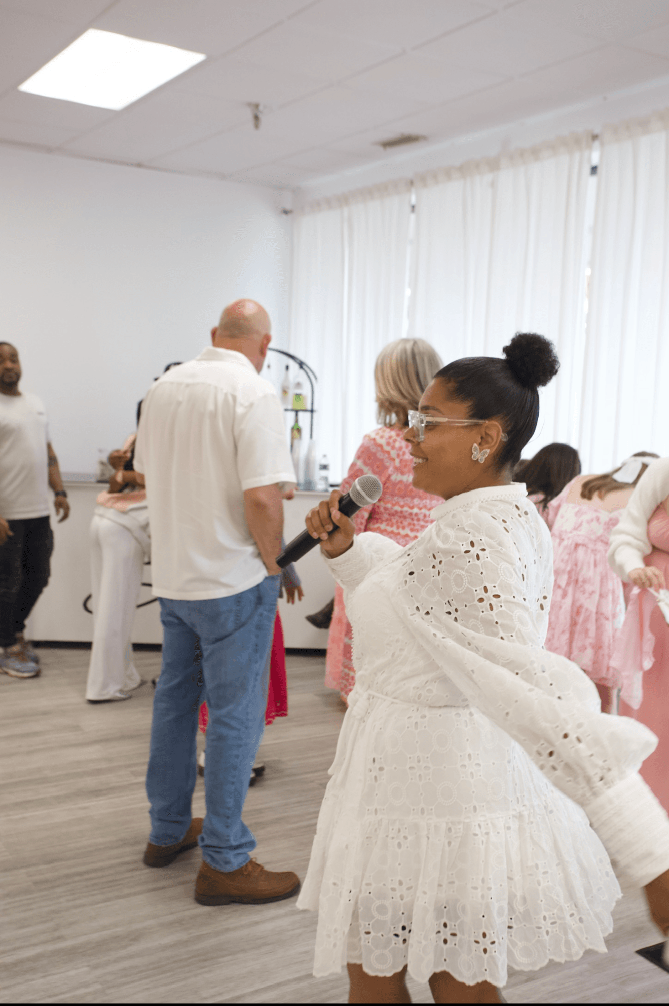 Smiling woman in a white eyelet dress holds a microphone at a social event.