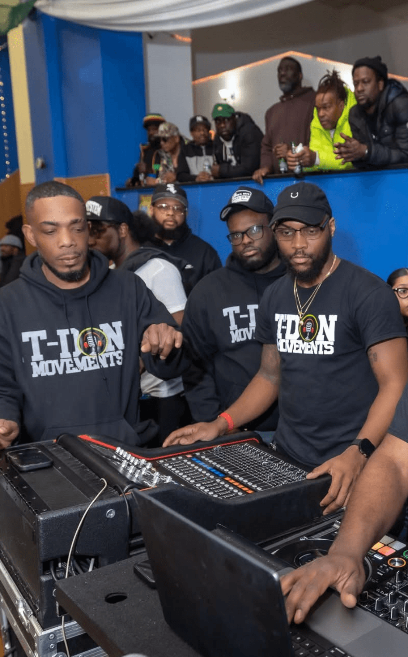 Men wearing T-ION MOVEMENTS shirts manage a professional audio mixing board at an event.