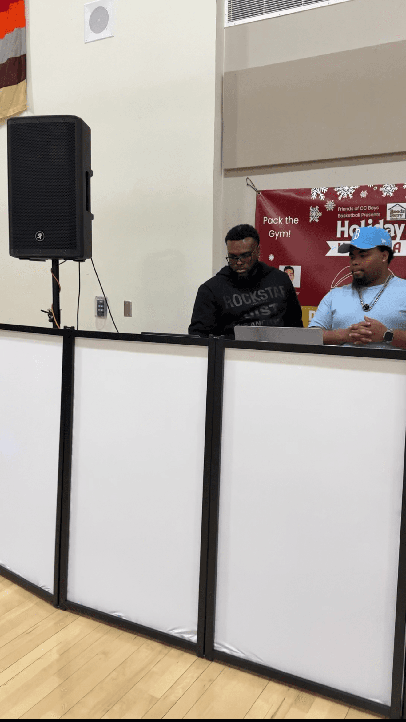 Two men behind a white DJ booth in a gym with a large black speaker.
