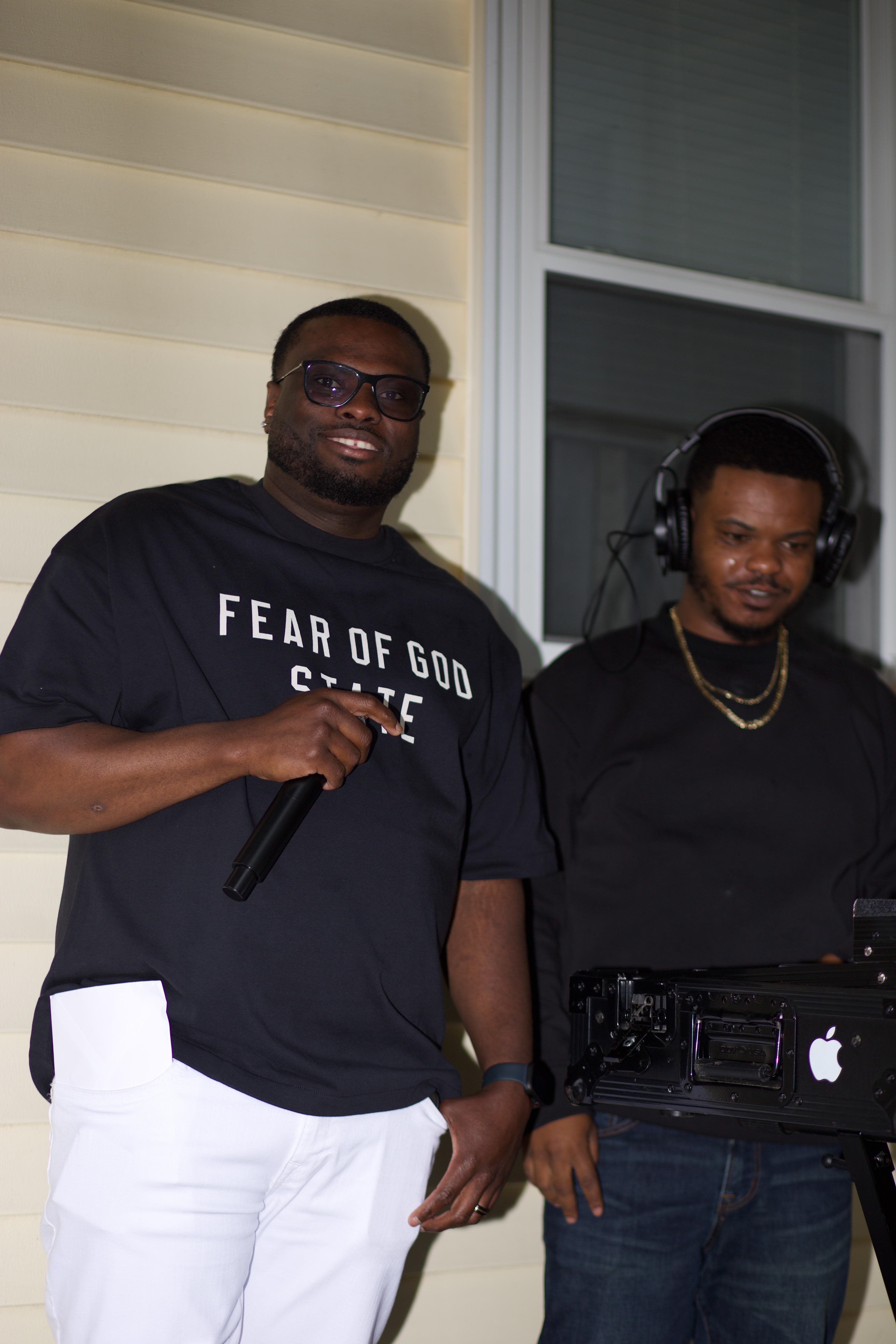 Smiling man holding a microphone stands beside a DJ wearing headphones at an outdoor event.